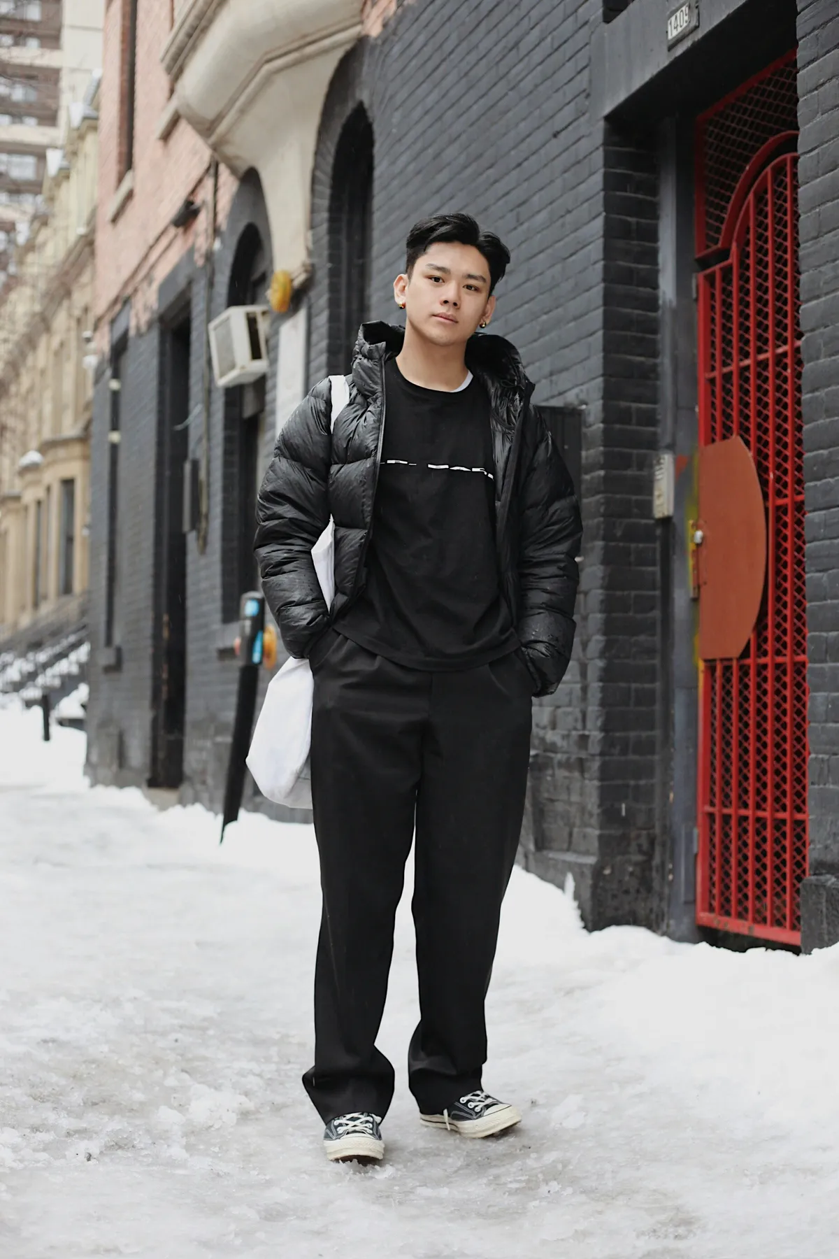 Man in a black puffer jacket, black trousers and sneakers on a snowy city sidewalk
